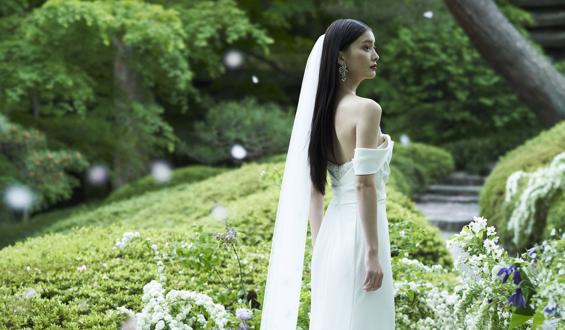 花と緑に囲まれた庭園で、白いウェディングドレスを着た女性の横顔の写真です。.