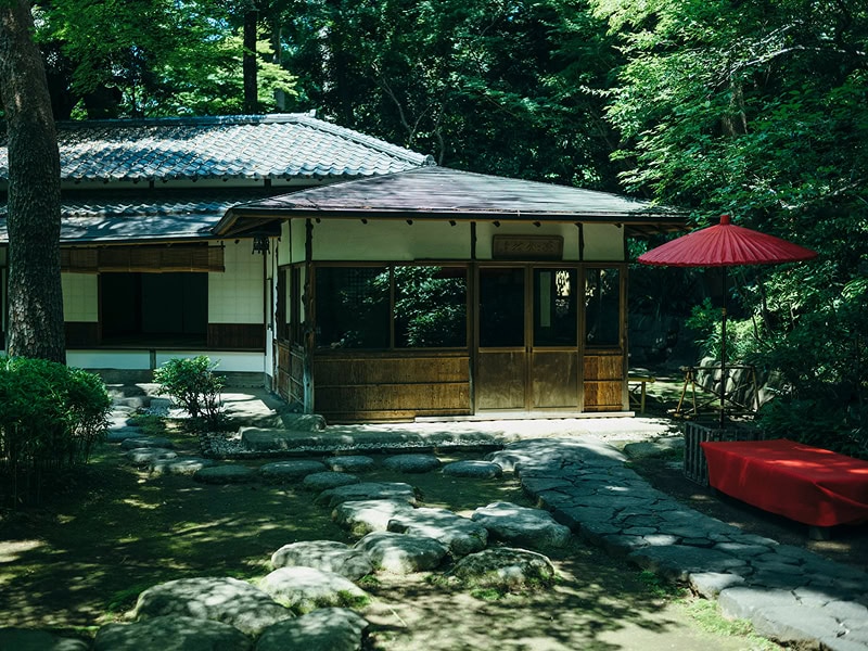日本庭園の伝統的な茶室と赤い傘、緑豊かな自然に囲まれた静かな風景.